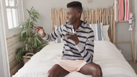 Happy-african-american-man-wearing-shorts-sitting-on-bed-talking-on-smartphone,-slow-motion