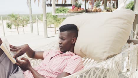 Happy-african-american-man-sitting-in-hammock-reading-book-on-sunny-beach,-slow-motion