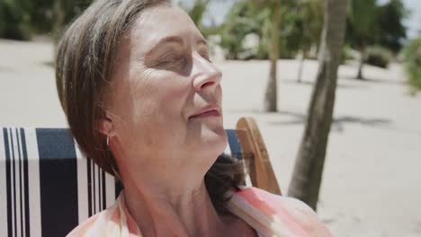 Smiling-senior-caucasian-woman-sitting-in-deckchair-on-sunny-beach-with-eyes-closed,-in-slow-motion