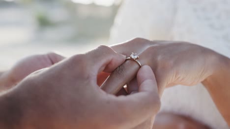 Hands-of-diverse-groom-putting-wedding-ring-on-finger-of-bride-at-beach-wedding,-in-slow-motion