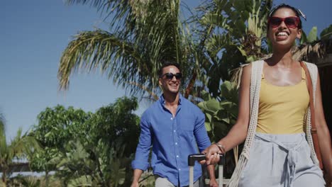 Happy-diverse-couple-in-sunglasses-walking-with-suitcases-on-sunny-beach,-in-slow-motion