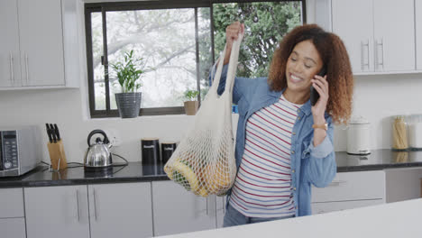Happy-biracial-woman-talking-on-phone-putting-down-bags-of-grocery-shopping-in-kitchen,-slow-motion