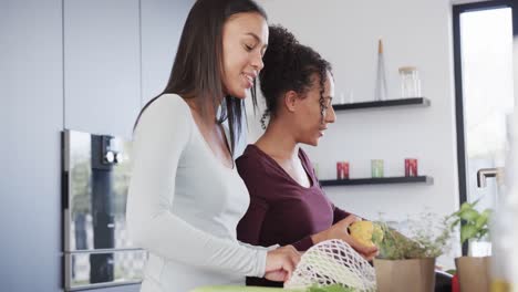 Happy-biracial-lesbian-couple-unpacking-grocery-shopping-and-talking-in-kitchen,-in-slow-motion