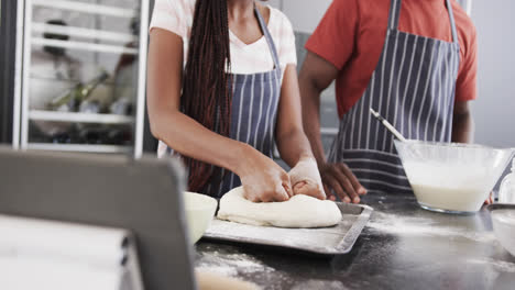 Happy-african-american-couple-in-aprons-baking-using-tablet,-kneading-dough-in-kitchen,-slow-motion