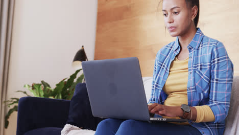 Focused-biracial-woman-sitting-on-couch-using-laptop-in-living-room,-in-slow-motion