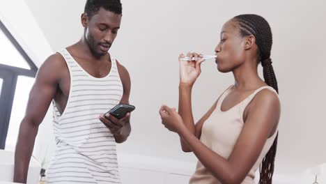 African-american-couple-brushing-teeth,-using-smartphone-and-talking-in-bathroom,-slow-motion