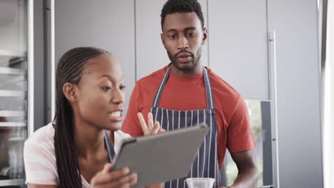 Happy-african-american-couple-in-aprons-baking-together-using-tablet-in-kitchen,-slow-motion