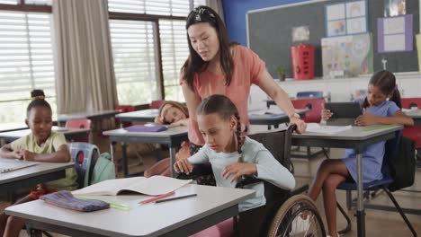 Diverse-female-teacher-teaching-schoolgirl-in-wheelchair-reading-in-classroom-at-elementary-school