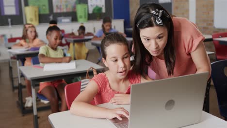 Happy-diverse-female-teacher-teaching-schoolgirls-using-laptop-in-classroom-at-elementary-school