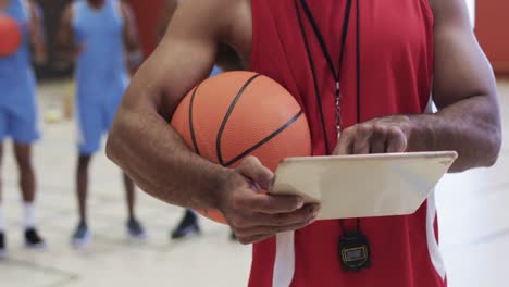 Midsection-of-biracial-male-basketball-coach-using-tablet-at-indoor-court,-in-slow-motion