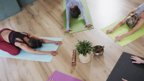 Multiracial-women-practicing-child-pose-around-plant-and-singing-bowl-in-yoga-studio