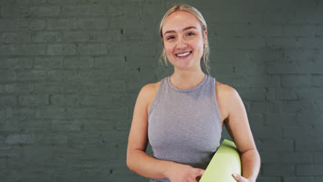 Smiling-caucasian-young-woman-holding-mat-in-front-of-grey-brick-wall-in-yoga-studio