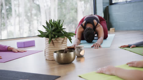Diverse-women-practicing-child-yoga-pose-around-plant-and-singing-bowl-in-studio