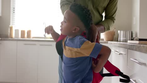 African-american-grandfather-giving-schoolbag-to-grandson-in-kitchen-before-school,-slow-motion