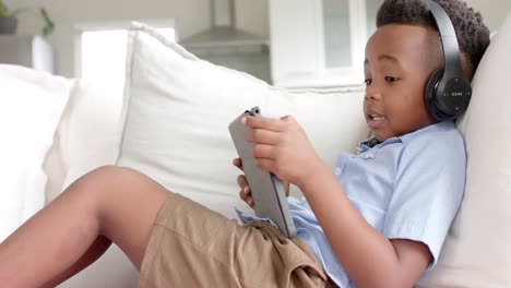 Happy-african-american-boy-in-headphones-lying-on-sofa-using-tablet-at-home,-slow-motion