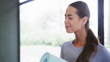 Smiling-biracial-young-woman-holding-mat-while-standing-against-window-in-yoga-studio