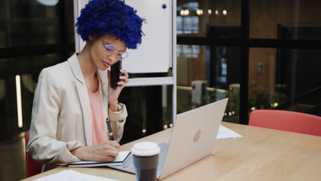 Biracial-businesswoman-with-blue-afro-talking-on-smartphone-and-using-laptop-at-desk,-slow-motion