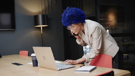 Biracial-businesswoman-with-blue-afro-talking-on-smartphone-and-using-laptop-at-desk,-slow-motion