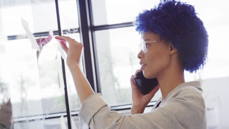 Biracial-casual-businesswoman-making-notes-on-glass-wall-and-talking-on-smartphone,-slow-motion