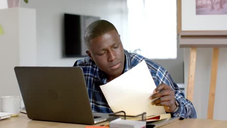 Afroamerikanischer-Mann-Sitzt-Mit-Laptop-Am-Tisch-Und-Hält-Dokumente-Zu-Hause,-Zeitlupe