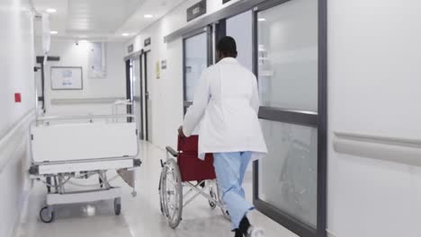 Afroamerikanischer-Männlicher-Arzt,-Der-Mit-Rollstuhl-Im-Krankenhausflur-Läuft,-Zeitlupe