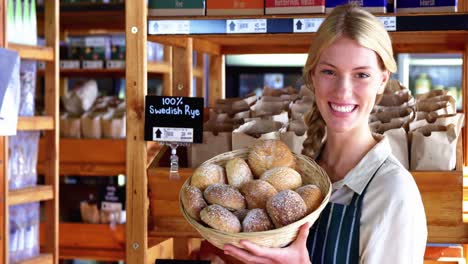 Smiling-female-staff-holding-basket-of-sesame-breads-at-bread-counter