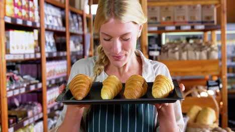 Smiling-female-staff-holding-tray-of-croissants-in-supermarket