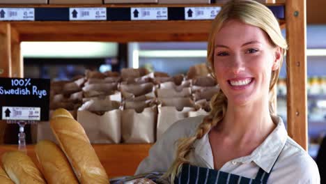 Smiling-female-staff-holding-basket-of-bread-at-bread-counter