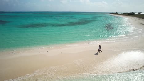 Drone-shot-woman-walk-backlight-sunset-on-white-sand-beach,-sea-water-splash-and-crash-on-sandbar