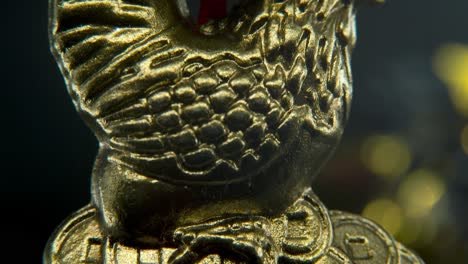 Chinese-New-Year-traditional-golden-chicken-zodiac-statue,-hanging-gold-rooster-sitting-on-money-coins-Asian-symbol,-glowing-background-with-blurry-depth-of-field,-cinematic-macro-tilt-up-4K-video