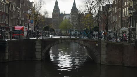 Aguas-Tranquilas-Del-Canal-Con-Panorámica-Hacia-Arriba-Para-Revelar-Un-Pequeño-Puente-Con-Un-Ciclista-Pasando-Por-Encima-En-Amsterdam