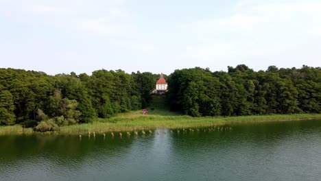 Old-building-next-to-a-green-natural-lake-surrounded-by-a-forest-in-Brandenburg,-Germany