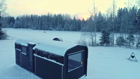 Trailer-and-sauna-by-a-frozen-lake-and-frosty-forest-in-the-countryside---aerial-flyover
