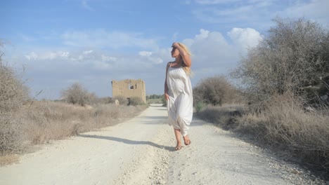 Wide-shot-of-a-woman-in-a-white-dress-searches-for-something-down-a-deserted-dirt-path-with-ruins-in-the-background