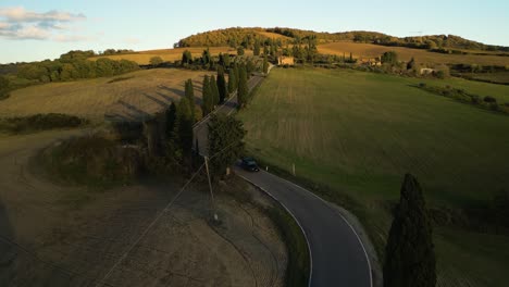 Cars-drive-along-winding-asphalt-road-across-farmland-fields-at-golden-hour-in-Tuscany