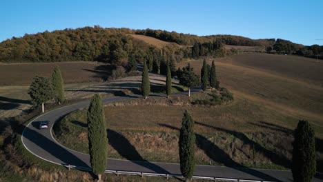 SUV-drives-up-along-winding-road-passing-across-Tuscan-landscape,-aerial-dolly