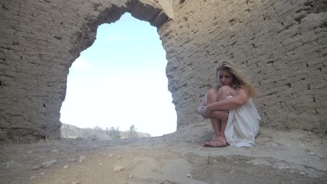 A-woman-in-a-white-dress-huddles-alone-under-a-deserted-ruin