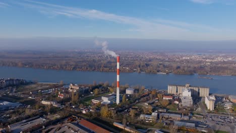 Aerial-footage-ascending-and-focusing-on-a-chimney-smokestack-in-the-city-of-Belgrade,-Serbia-with-the-Danube-River-in-the-background