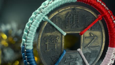 Verträumte-Nahaufnahme-Einer-Alten-Chinesischen-Glücksmünze,-Asiatische-Neujahrsdekoration,-Golden-Glänzender,-Unscharfer-Hintergrund,-Quadratisches-Loch,-Bunte-Schnüre,-Kulturelles-Feiertagssymbol,-Glatte-Pfanne-Nach-Links,-4K-Video