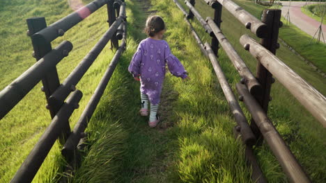 Following-shot-of-Playful-Little-Girl-Walking-Up-the-hill-on-Grassy-Trail-Way-Along-Wooden-Logs-Fence-in-slow-motion