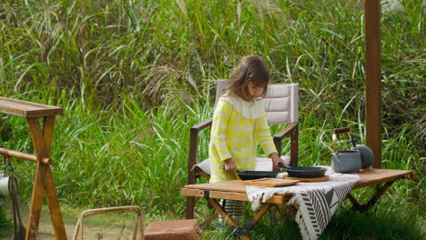 Curious-Little-Girl-Playing-with-Frying-Pans-Standing-by-Wooden-Table-at-Camping-Ground-Outdoors-in-Country