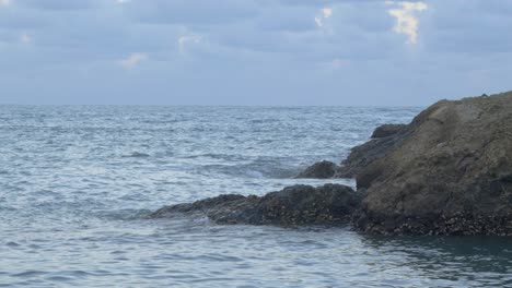 Gentle-wave-swells-breaking-on-rocky-sea-shore-with-calm-gray-clouds-floating-in-sky,-filmed-as-stationary-shot