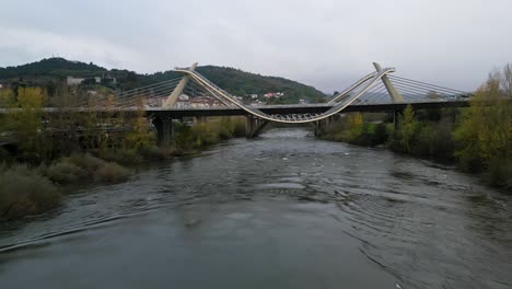 Millennium-Bridge-over-the-Miño-River-in-Ourense,-Galicia,-Spain,-drone-pullback-above-autumnal-trees-and-cloudy-grey-sky