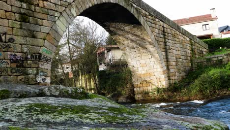 Angled-view-of-sunlight-on-brick-bridge-with-mossy-lichen-covered-rock-by-lonia-river-in-Ourense