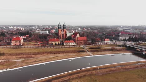 Drone-reverse-dolly-from-red-Cathedral-in-Poznan,-Poland-with-snow-covered-riverbank