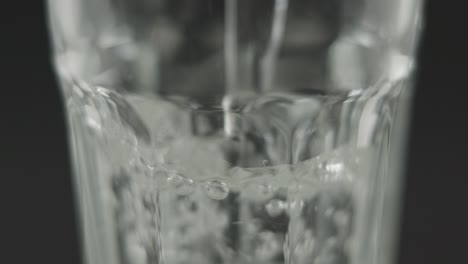 Slowmotion-closeup-of-still-water-pouring-into-clear-glass-in-front-of-dark-background