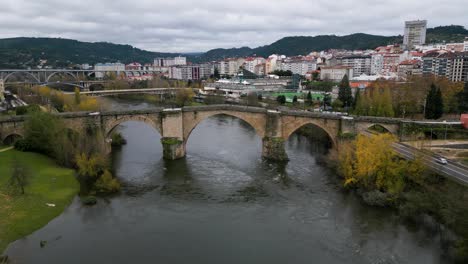 Drone-pulls-back-as-tourists-walk-across-Ourense-Roman-Bridge-on-Miño-River-in-Ourense,-Galicia,-Spain