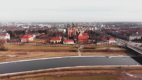 Vibrant-red-brick-Cathedral-of-Poznan,-Poland-looks-out-over-river-off-highway,-aerial