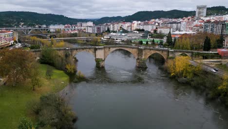 Drone-descends-to-river-with-reflection-of-Ourense-Roman-Bridge-on-Miño-River-in-Ourense,-Galicia,-Spain