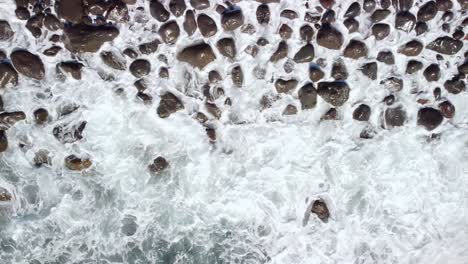 Top-down-background-view-of-waves-foam-splashing-on-pebbles-rock-coast,-Tenerife